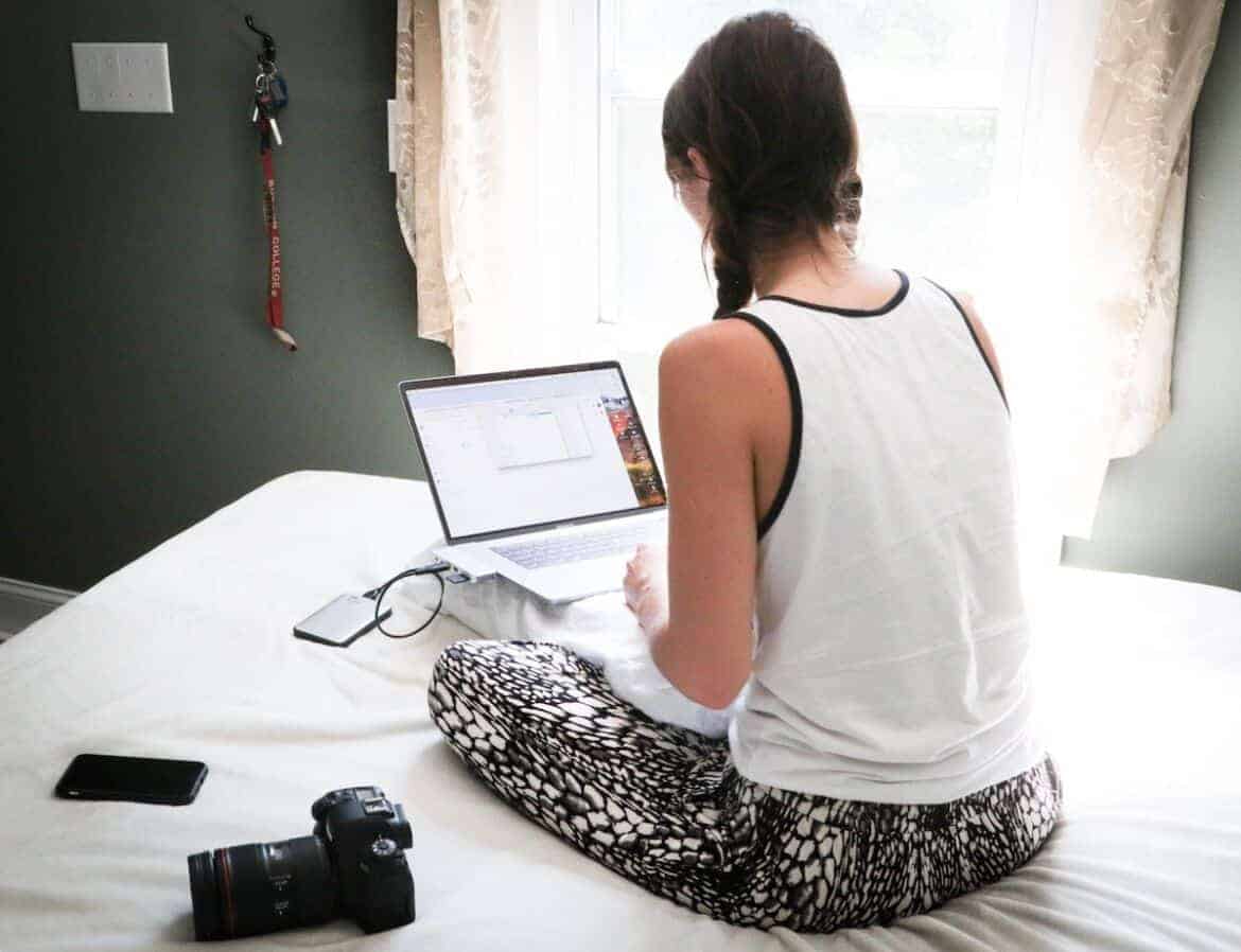 woman in white tank top sitting on bed in front of laptop computer